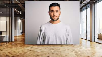 Young handsome man wearing casual t-shirt standing over isolated white background with serious expression on face. Simple and natural looking at the camera. Wall mural