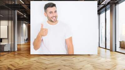 Young handsome man wearing casual white t-shirt over white isolated background doing happy thumbs up gesture with hand. Approving expression looking at the camera with showing success. Wall mural