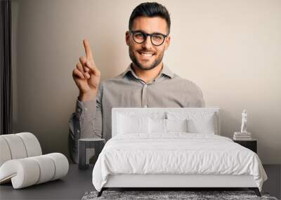 Young handsome man wearing elegant shirt and glasses over isolated white background showing and pointing up with finger number one while smiling confident and happy. Wall mural