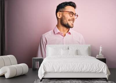 Young handsome man wearing elegant shirt and glasses standing over pink background looking away to side with smile on face, natural expression. Laughing confident. Wall mural