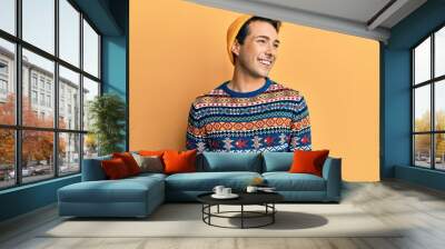 Young handsome man wearing wool hat and colorful sweater looking away to side with smile on face, natural expression. laughing confident. Wall mural