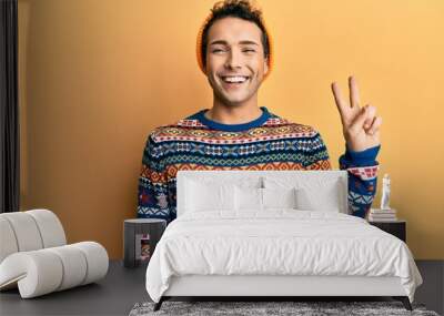 Young handsome man wearing wool hat and colorful sweater smiling with happy face winking at the camera doing victory sign. number two. Wall mural