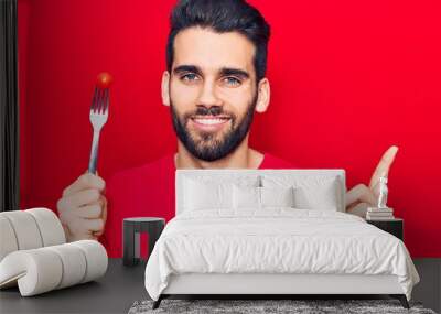 Young handsome man with beard holding fork with tomato smiling happy pointing with hand and finger to the side Wall mural