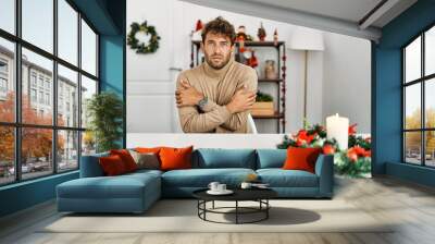 Young handsome man with beard sitting on the table by christmas decoration shaking and freezing for winter cold with sad and shock expression on face Wall mural