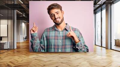 Young handsome man with beard wearing casual shirt standing over pink background smiling and looking at the camera pointing with two hands and fingers to the side. Wall mural