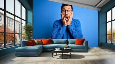 Young handsome man with beard wearing casual sweater and glasses over blue background afraid and shocked, surprise and amazed expression with hands on face Wall mural