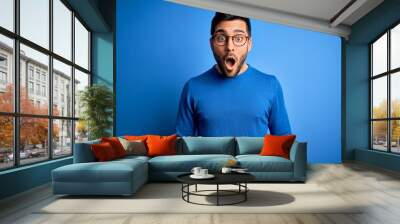 Young handsome man with beard wearing casual sweater and glasses over blue background afraid and shocked with surprise and amazed expression, fear and excited face. Wall mural