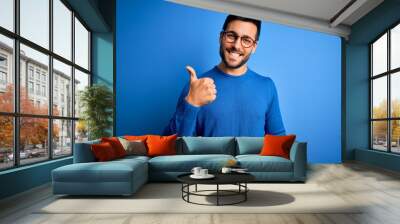 Young handsome man with beard wearing casual sweater and glasses over blue background doing happy thumbs up gesture with hand. Approving expression looking at the camera showing success. Wall mural