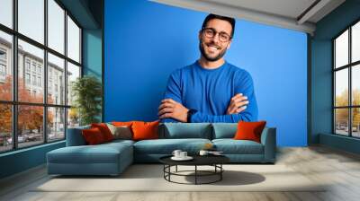 Young handsome man with beard wearing casual sweater and glasses over blue background happy face smiling with crossed arms looking at the camera. Positive person. Wall mural