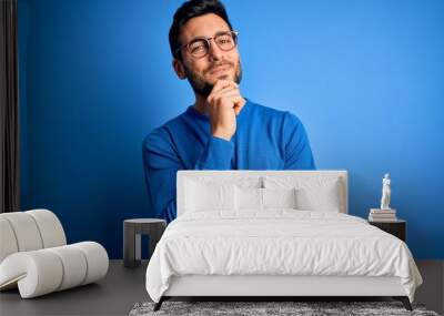 Young handsome man with beard wearing casual sweater and glasses over blue background looking confident at the camera smiling with crossed arms and hand raised on chin. Thinking positive. Wall mural