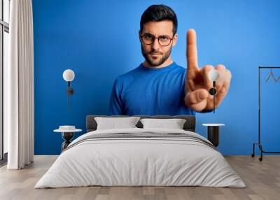 Young handsome man with beard wearing casual sweater and glasses over blue background Pointing with finger up and angry expression, showing no gesture Wall mural