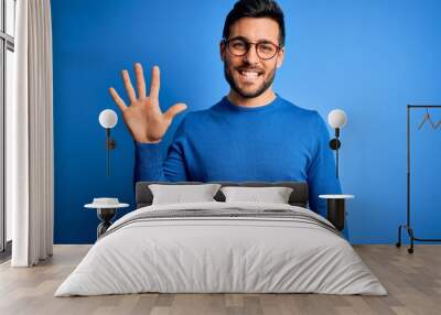 Young handsome man with beard wearing casual sweater and glasses over blue background showing and pointing up with fingers number five while smiling confident and happy. Wall mural