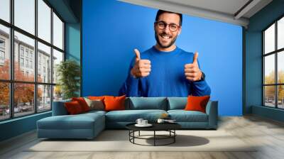 Young handsome man with beard wearing casual sweater and glasses over blue background success sign doing positive gesture with hand, thumbs up smiling and happy. Cheerful expression and winner Wall mural