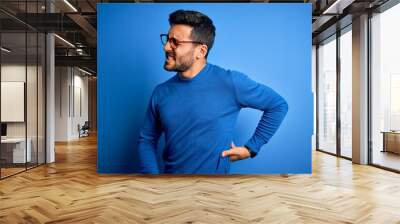 Young handsome man with beard wearing casual sweater and glasses over blue background Suffering of backache, touching back with hand, muscular pain Wall mural