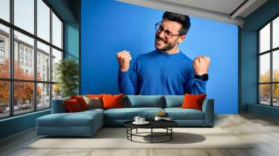 Young handsome man with beard wearing casual sweater and glasses over blue background very happy and excited doing winner gesture with arms raised, smiling and screaming for success. Celebration Wall mural