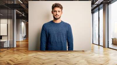 Young handsome man with beard wearing casual sweater standing over white background Relaxed with serious expression on face. Simple and natural looking at the camera. Wall mural