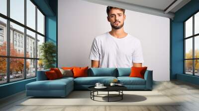 Young handsome man with beard wearing casual t-shirt standing over white background Relaxed with serious expression on face. Simple and natural looking at the camera. Wall mural