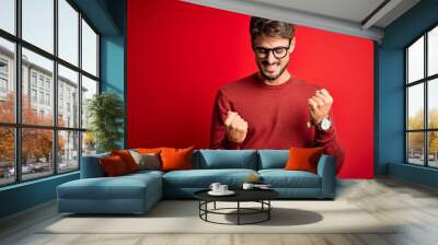 Young handsome man with beard wearing glasses and sweater standing over red background celebrating surprised and amazed for success with arms raised and eyes closed. Winner concept. Wall mural