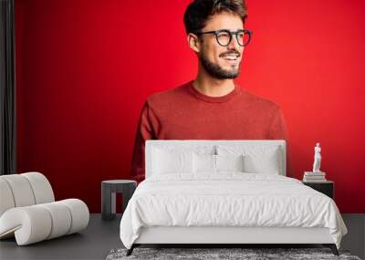 Young handsome man with beard wearing glasses and sweater standing over red background looking away to side with smile on face, natural expression. Laughing confident. Wall mural