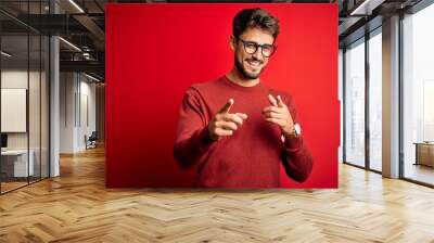 Young handsome man with beard wearing glasses and sweater standing over red background pointing fingers to camera with happy and funny face. Good energy and vibes. Wall mural