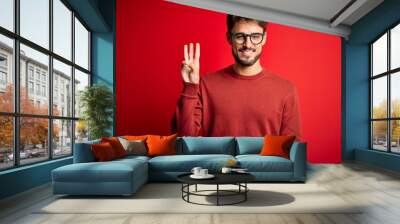 Young handsome man with beard wearing glasses and sweater standing over red background showing and pointing up with fingers number three while smiling confident and happy. Wall mural