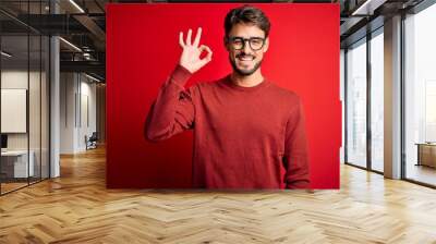 Young handsome man with beard wearing glasses and sweater standing over red background smiling positive doing ok sign with hand and fingers. Successful expression. Wall mural