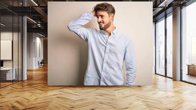 Young handsome man with beard wearing striped shirt standing over white background smiling confident touching hair with hand up gesture, posing attractive and fashionable Wall mural