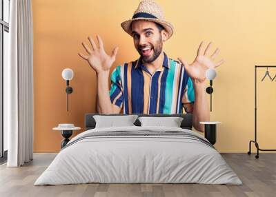 Young handsome man with beard wearing summer hat and shirt showing and pointing up with fingers number ten while smiling confident and happy. Wall mural