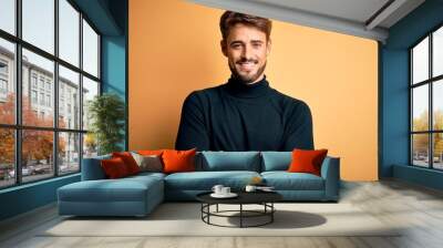 Young handsome man with beard wearing turtleneck sweater standing over yellow background happy face smiling with crossed arms looking at the camera. Positive person. Wall mural