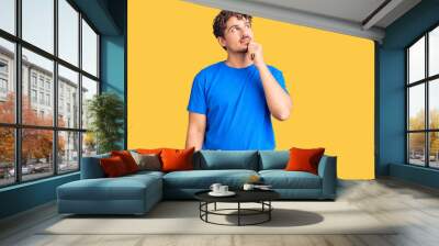 Young handsome man with curly hair wearing casual clothes with hand on chin thinking about question, pensive expression. smiling and thoughtful face. doubt concept. Wall mural