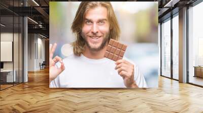 Young handsome man with long hair eating chocolate bar over isolated background doing ok sign with fingers, excellent symbol Wall mural