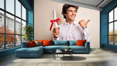 Young handsome student man holding diploma standing over isolated pink background pointing and showing with thumb up to the side with happy face smiling Wall mural