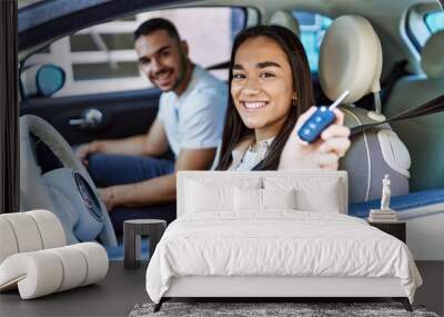 Young hispanic couple driving auto at the city. Girl smiling happy holding key of new car. Wall mural