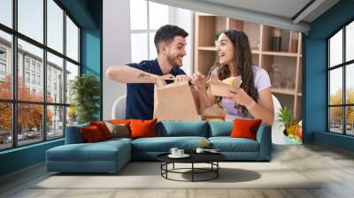 Young hispanic couple eating take away food sitting on table at home Wall mural