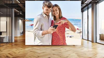 Young hispanic couple on vacation smiling happy using smartphone at the beach Wall mural