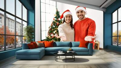 Young hispanic couple standing by christmas tree smiling cheerful offering palm hand giving assistance and acceptance. Wall mural