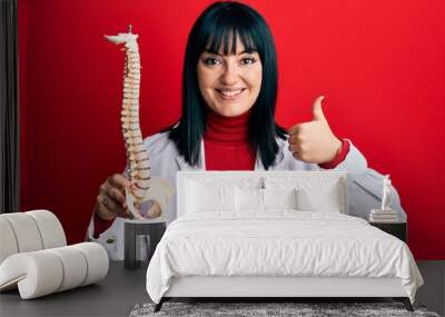 Young hispanic doctor woman holding anatomical model of spinal column smiling happy and positive, thumb up doing excellent and approval sign Wall mural