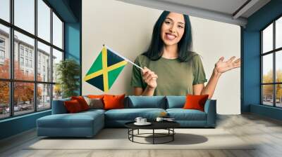 Young hispanic girl holding jamaica flag celebrating achievement with happy smile and winner expression with raised hand Wall mural