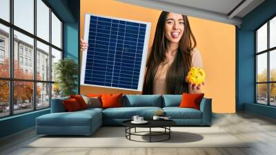 Young hispanic girl holding photovoltaic solar panel and piggy bank sticking tongue out happy with funny expression. Wall mural