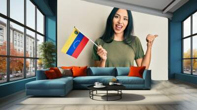 Young hispanic girl holding venezuelan flag pointing thumb up to the side smiling happy with open mouth Wall mural