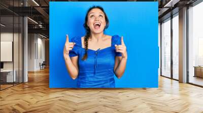 Young hispanic girl wearing casual clothes amazed and surprised looking up and pointing with fingers and raised arms. Wall mural