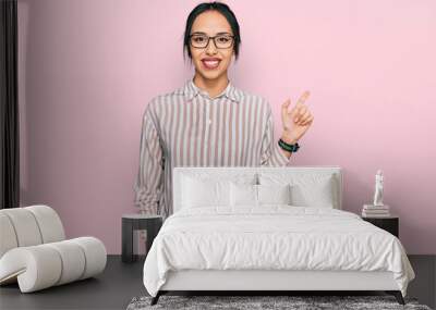 Young hispanic girl wearing casual clothes and glasses with a big smile on face, pointing with hand finger to the side looking at the camera. Wall mural