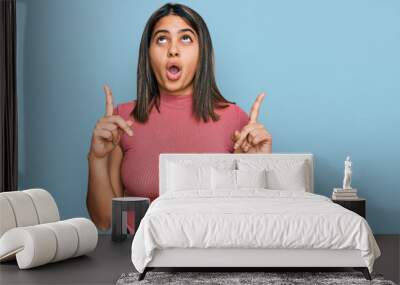 Young hispanic girl wearing casual t shirt amazed and surprised looking up and pointing with fingers and raised arms. Wall mural