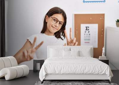 Young hispanic girl wearing glasses smiling looking to the camera showing fingers doing victory sign. number two. Wall mural