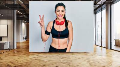 Young hispanic girl wearing gym clothes and using headphones showing and pointing up with fingers number two while smiling confident and happy. Wall mural