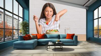 Young hispanic kid sitting on the table eating healthy salad very happy pointing with hand and finger Wall mural