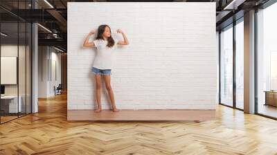 Young hispanic kid stading over white brick wall showing arms muscles smiling proud. Fitness concept. Wall mural