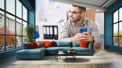 Young hispanic man business worker using laptop talking on smartphone at office Wall mural