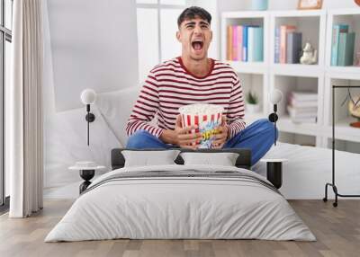 Young hispanic man eating popcorn sitting on the bed watching a movie angry and mad screaming frustrated and furious, shouting with anger looking up. Wall mural