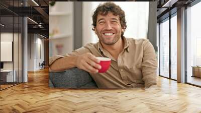 Young hispanic man holding cup of coffee sitting on sofa smiling at home Wall mural